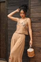Woman in a beige embroidered top and pants standing against a wooden wall.