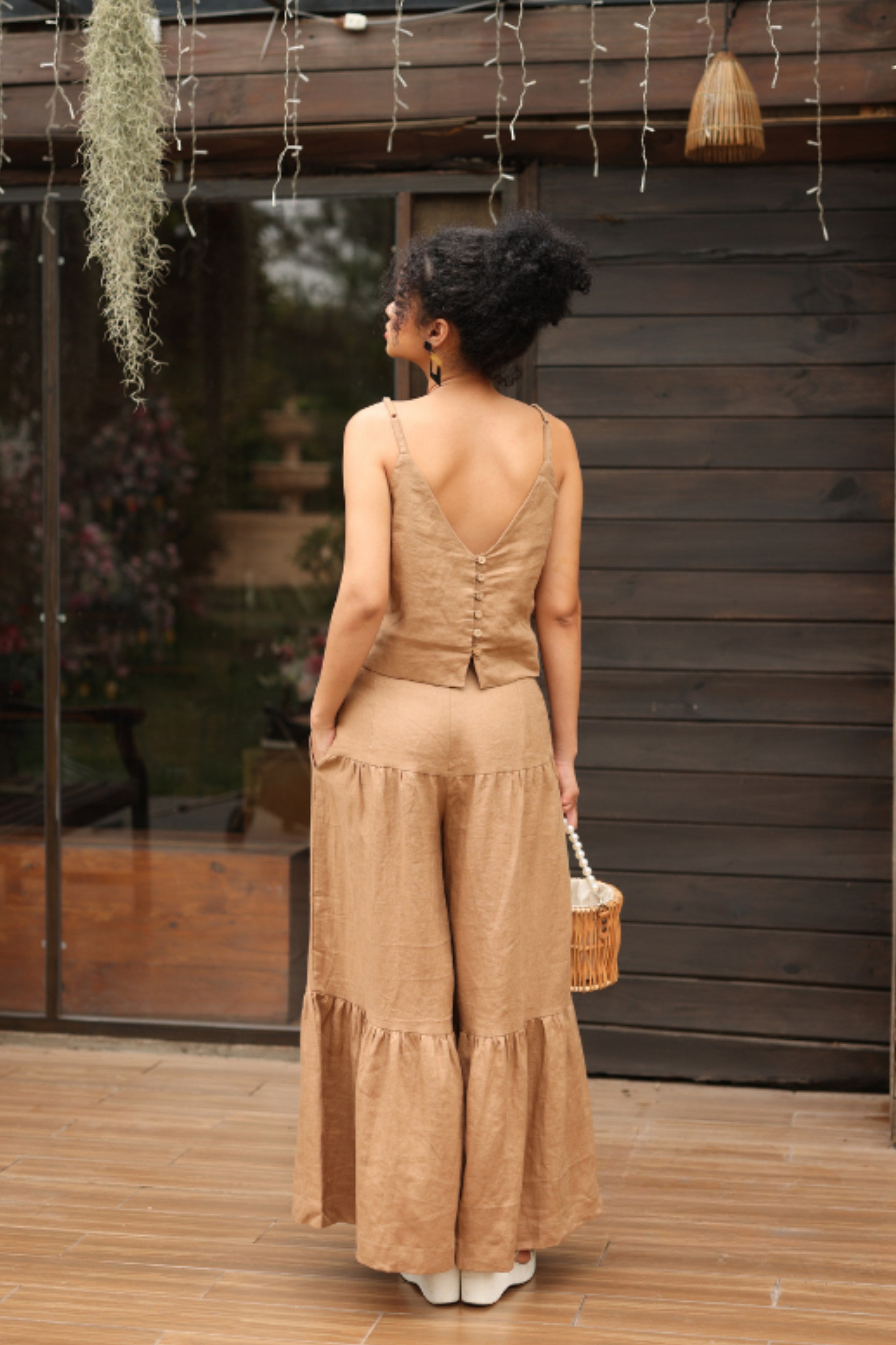 Woman in a beige outfit standing on a wooden deck with a wooden wall and plants in the background.