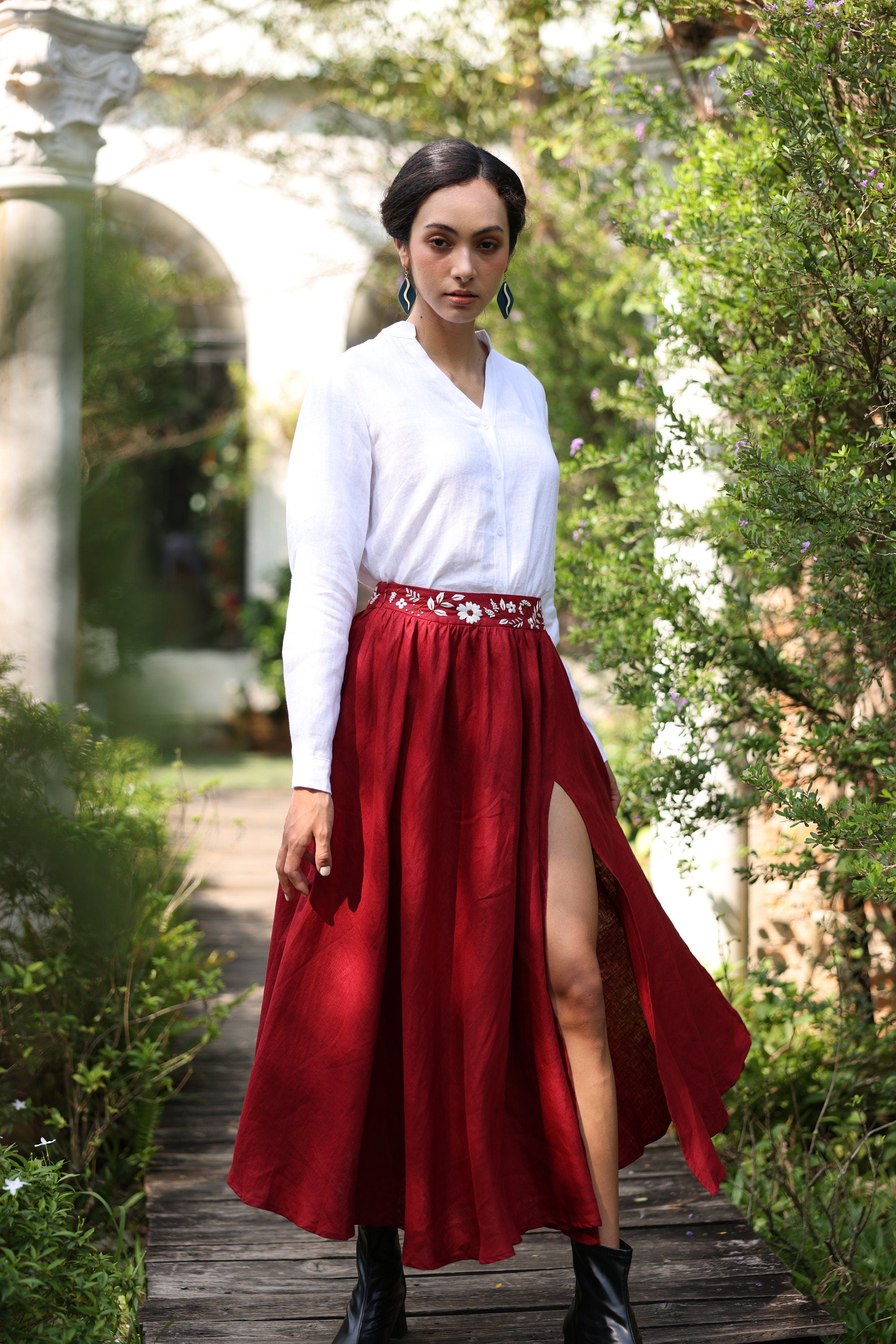 Woman in a white blouse and red skirt standing outdoors with greenery around