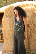 Woman standing in front of hay bales wearing a sleeveless top with floral embroidery and pants.