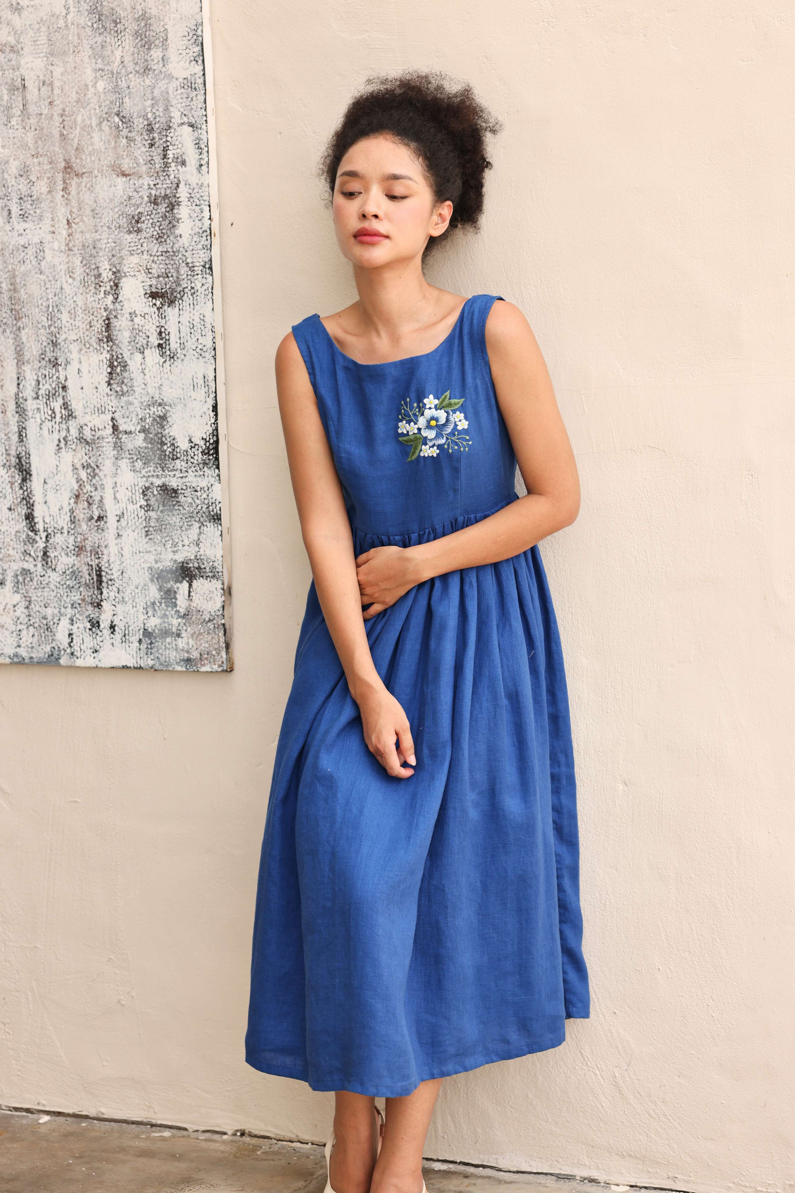 Woman wearing a blue dress with floral embroidery against a neutral wall.