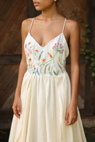 Woman wearing an ivory dress with floral embroidery against a wooden wall.