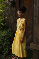 Woman in a yellow dress standing against a wooden wall with greenery.