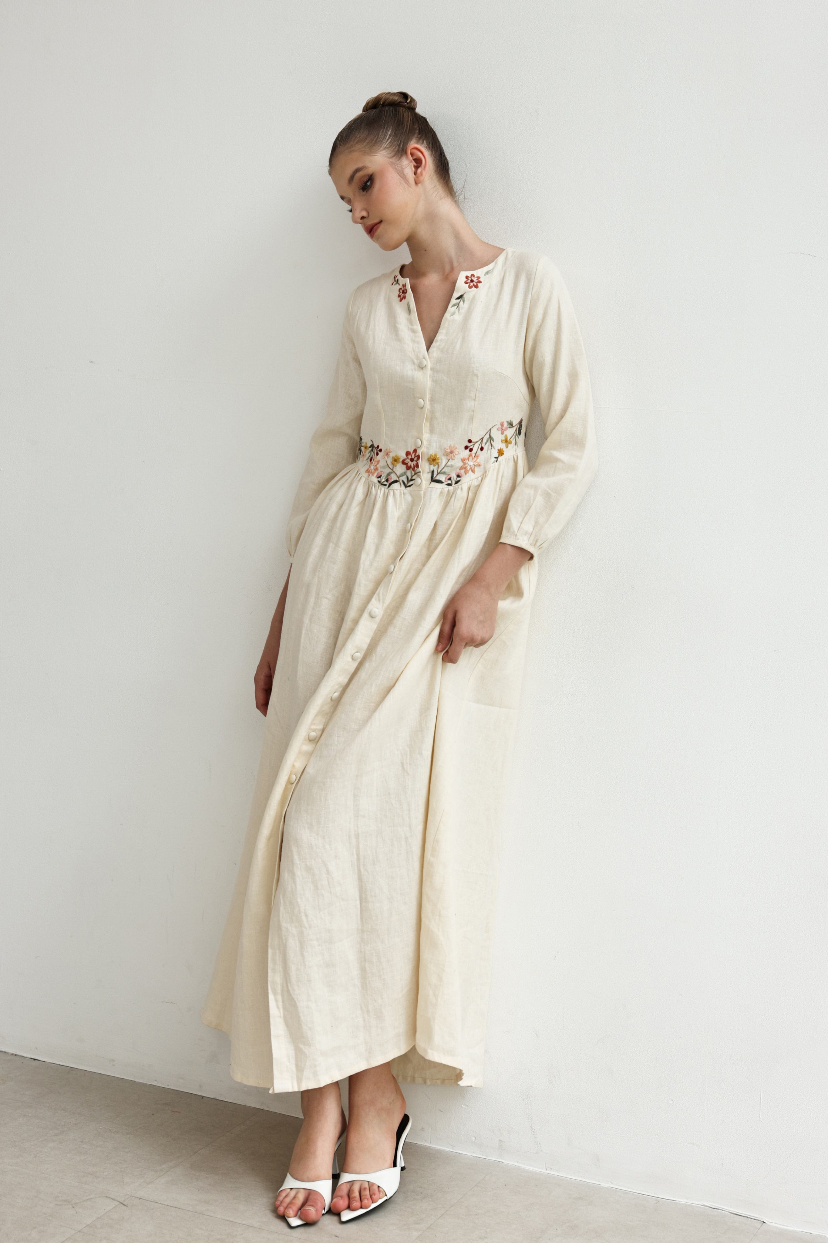 Woman wearing a long, light-colored dress with floral details on a plain background