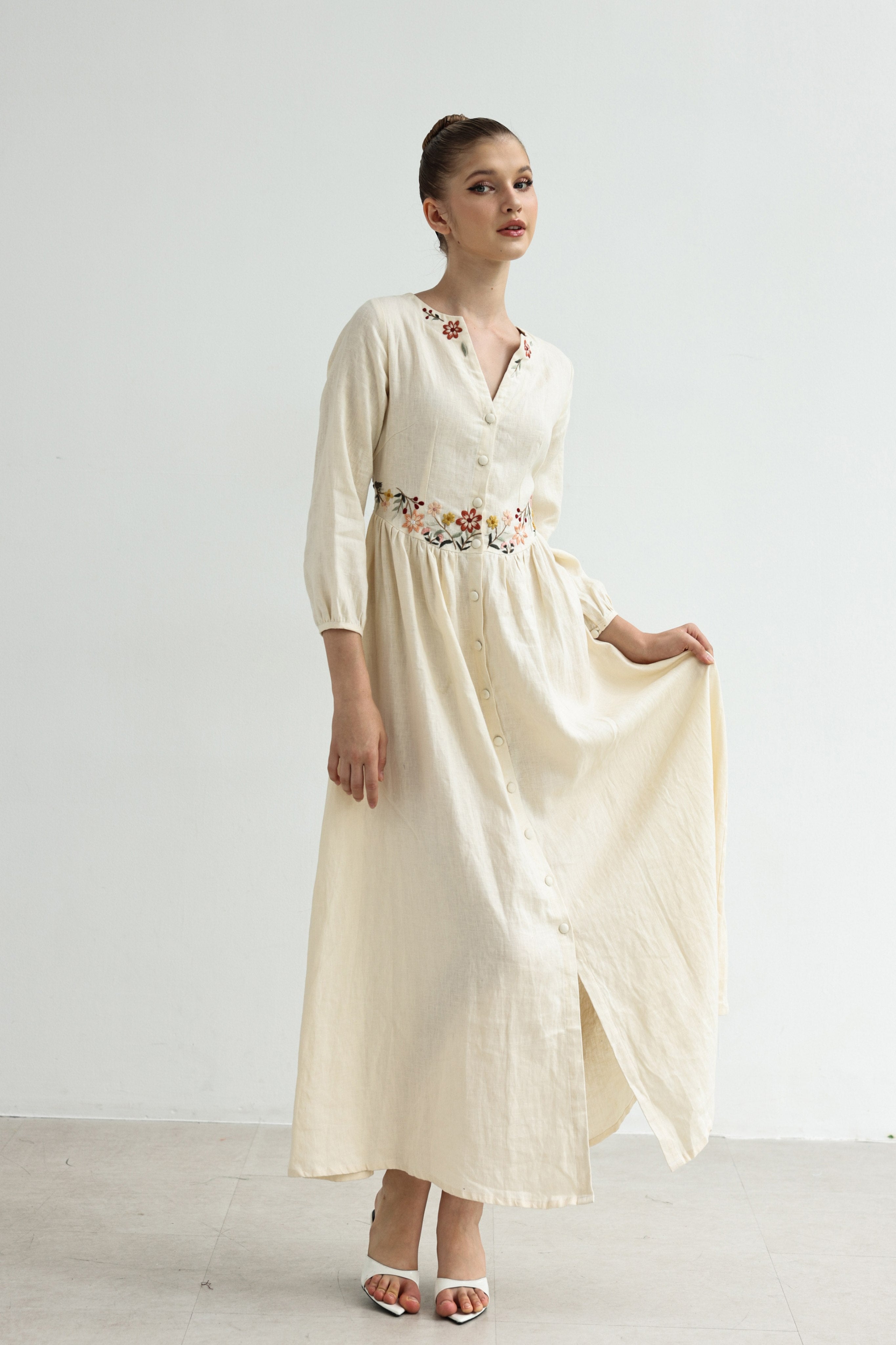 Woman wearing a long, light-colored dress with floral embellishments on a plain background