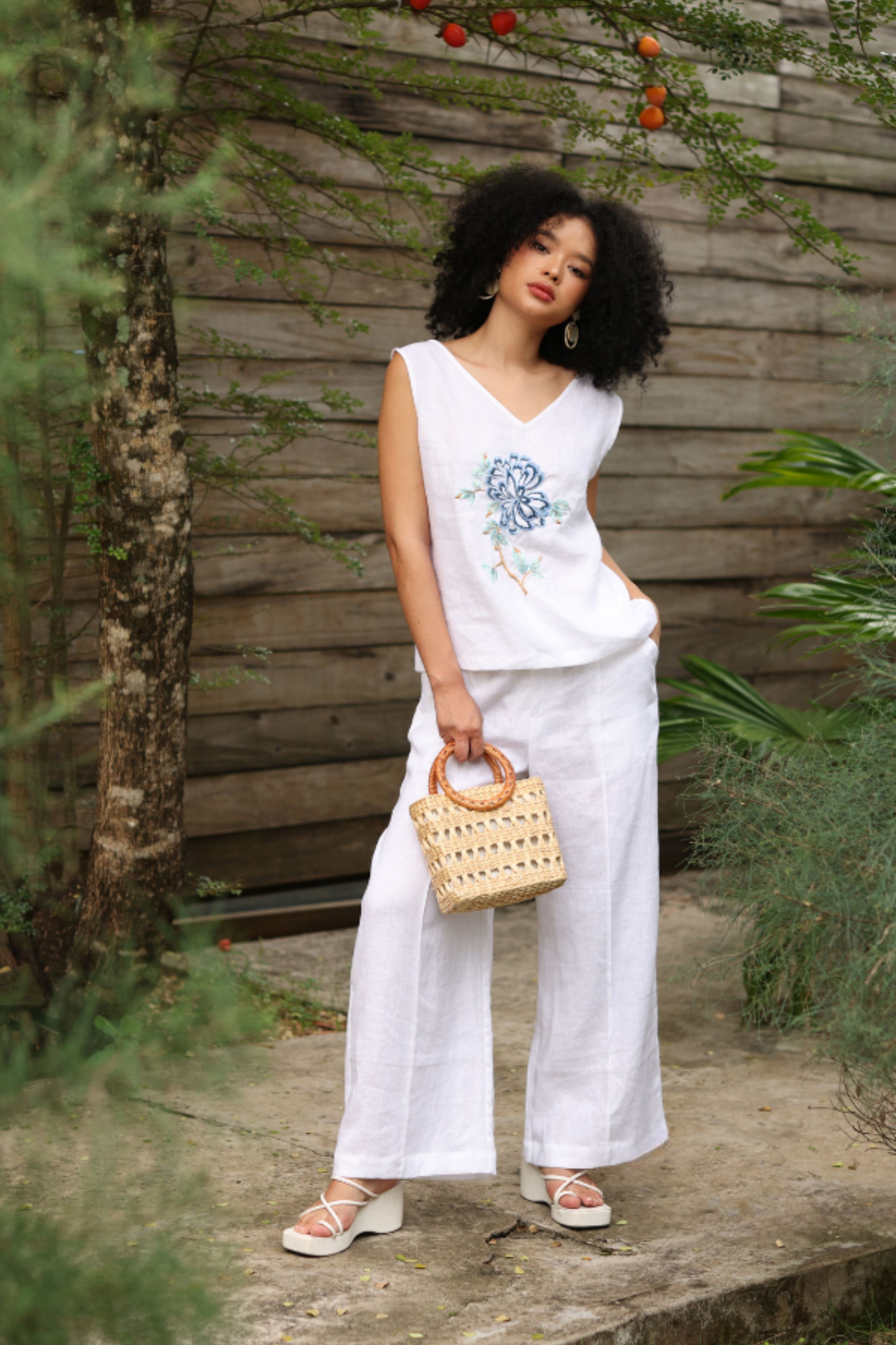 Woman in a white outfit with floral design standing outdoors against a wooden wall.