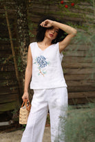 Woman wearing a white sleeveless top with floral design and white pants, standing outdoors.