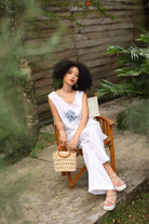 Woman in a white outfit sitting on a wooden chair outdoors with plants around.