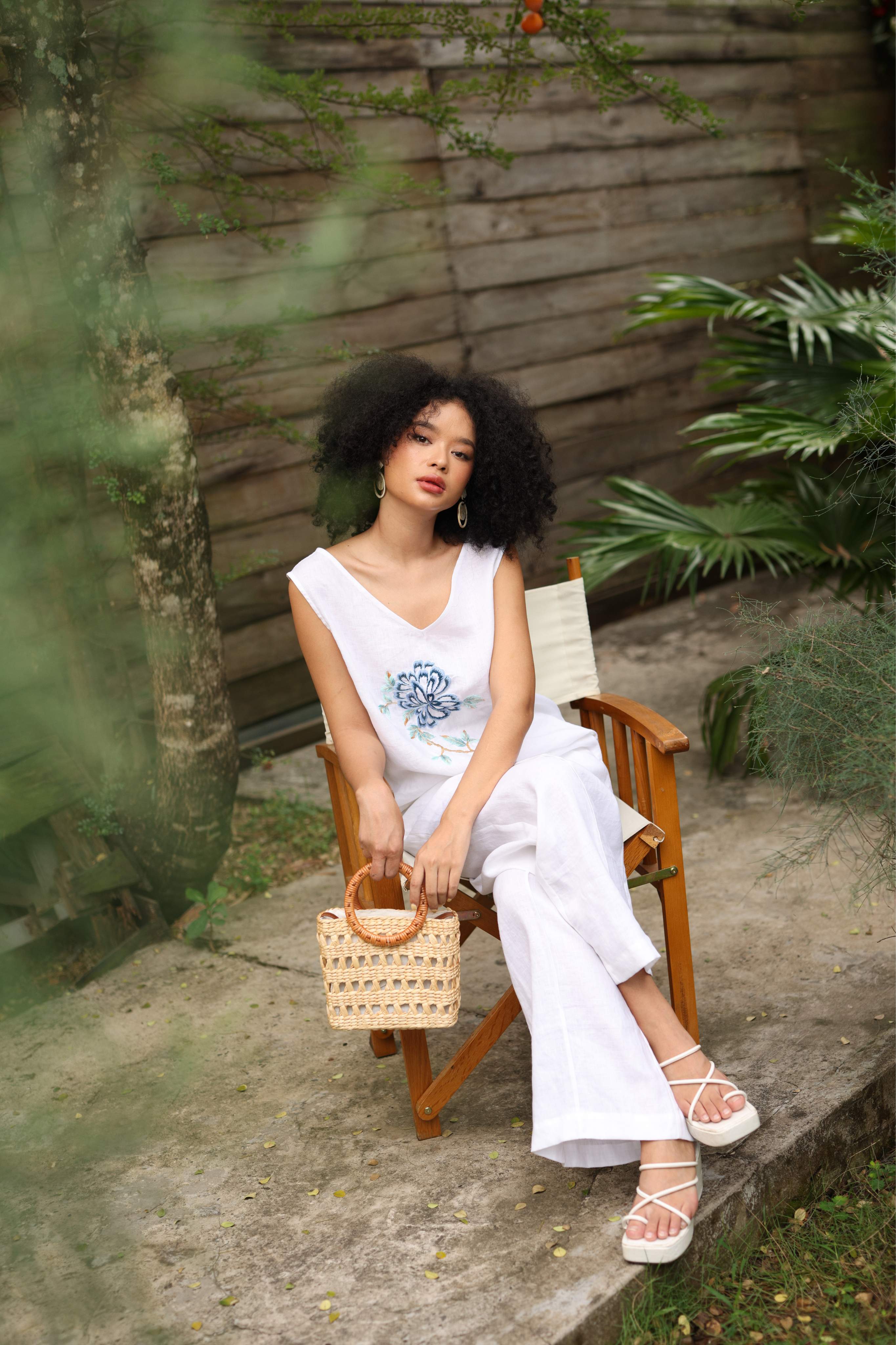 Woman in a white outfit sitting on a wooden chair outdoors with plants around.