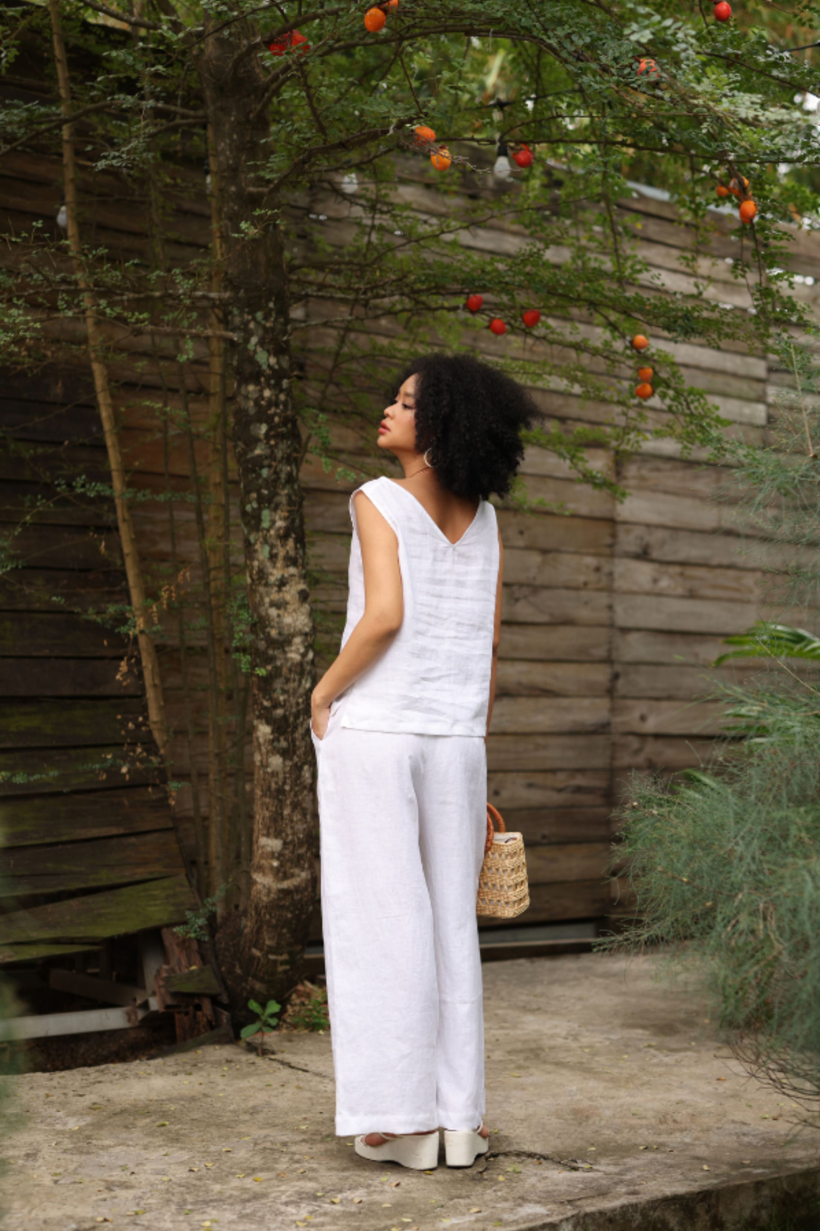 Woman in a white outfit standing in a garden with wooden walls and hanging fruit.