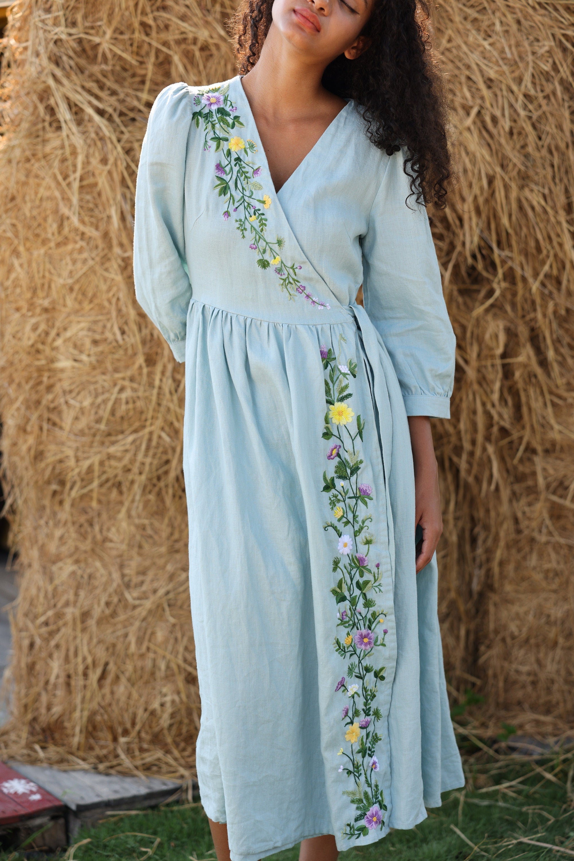 Woman wearing a light blue dress with floral embroidery against a straw bale background