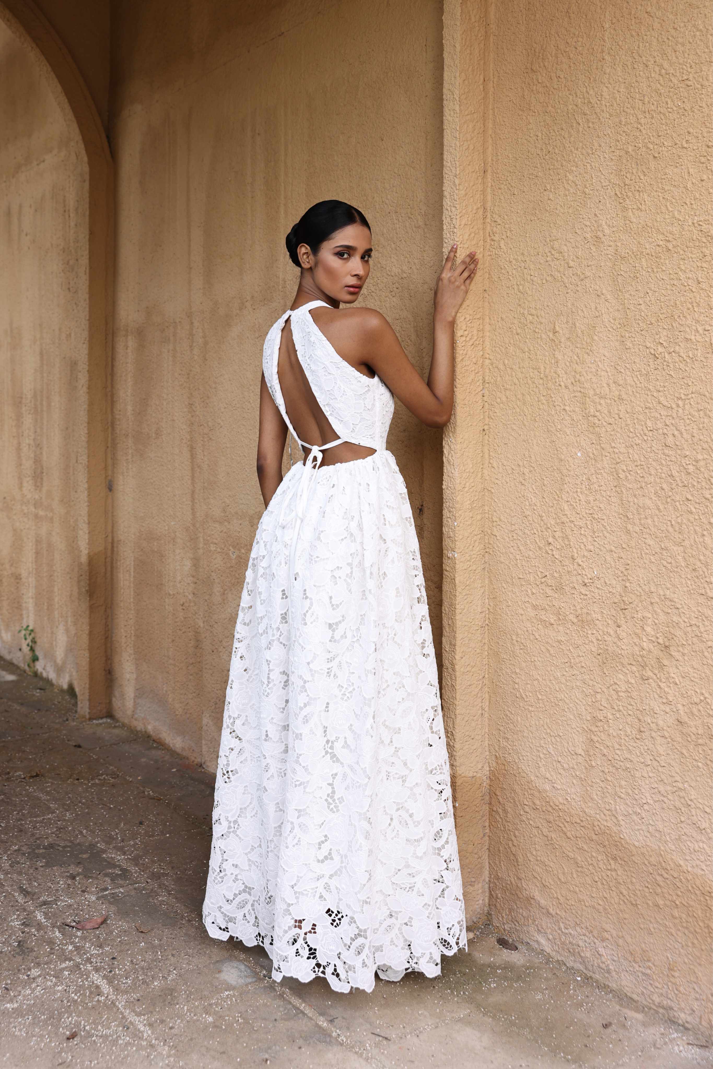 Woman in a white lace dress standing against a beige wall.