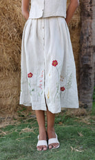 Person wearing a light-colored skirt with floral embroidery standing in a natural setting.