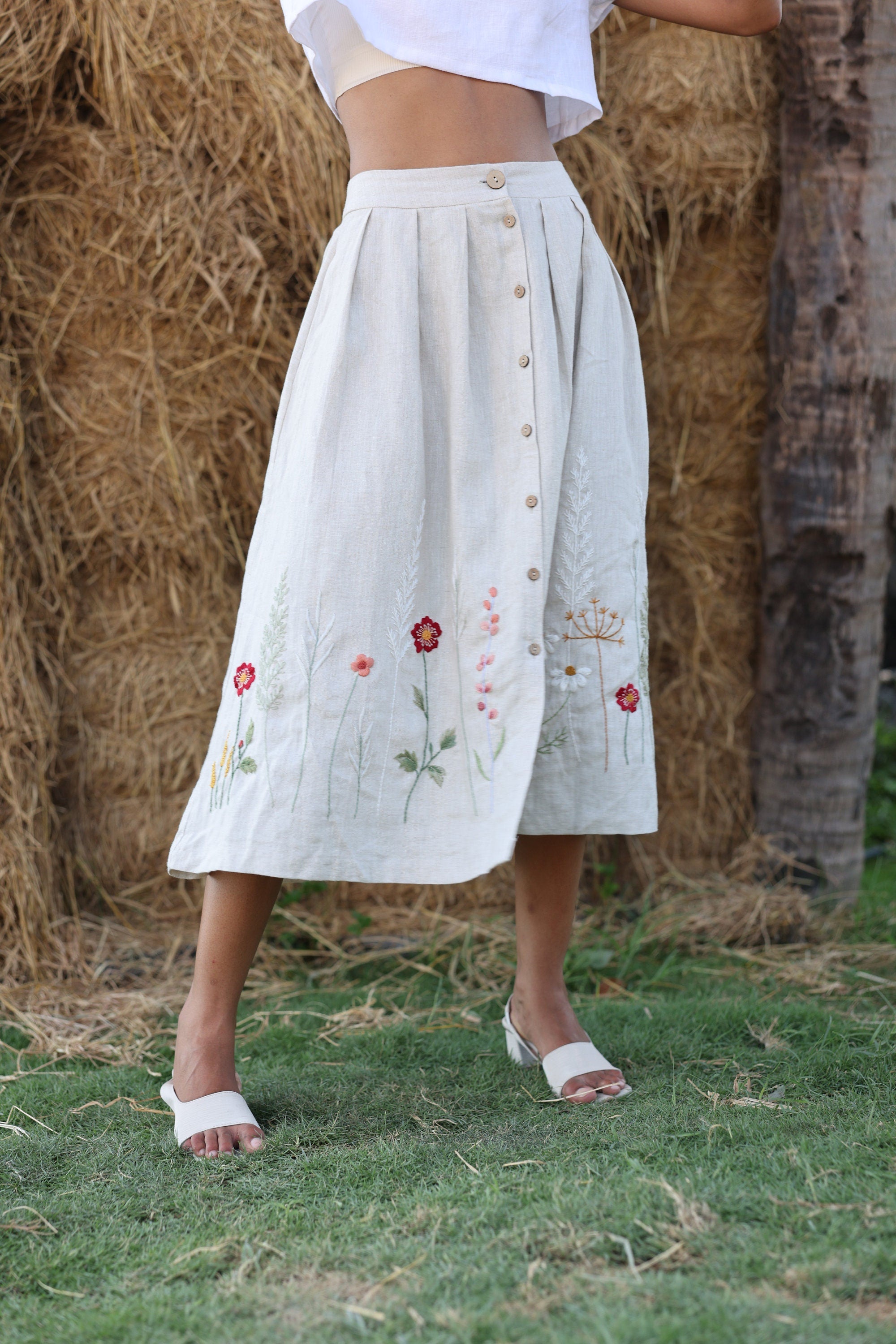 Person wearing a white skirt with floral embroidery standing in front of hay bales.
