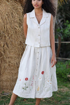 Woman wearing a white sleeveless top and skirt with floral embroidery, standing in front of hay bales.