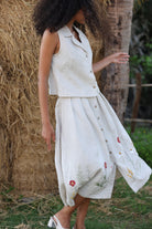 Woman wearing a white sleeveless top and skirt with floral embroidery standing near a tree and hay bale.