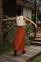Person in a white sleeveless top and rust-colored skirt standing on a wooden path with greenery and a building in the background.