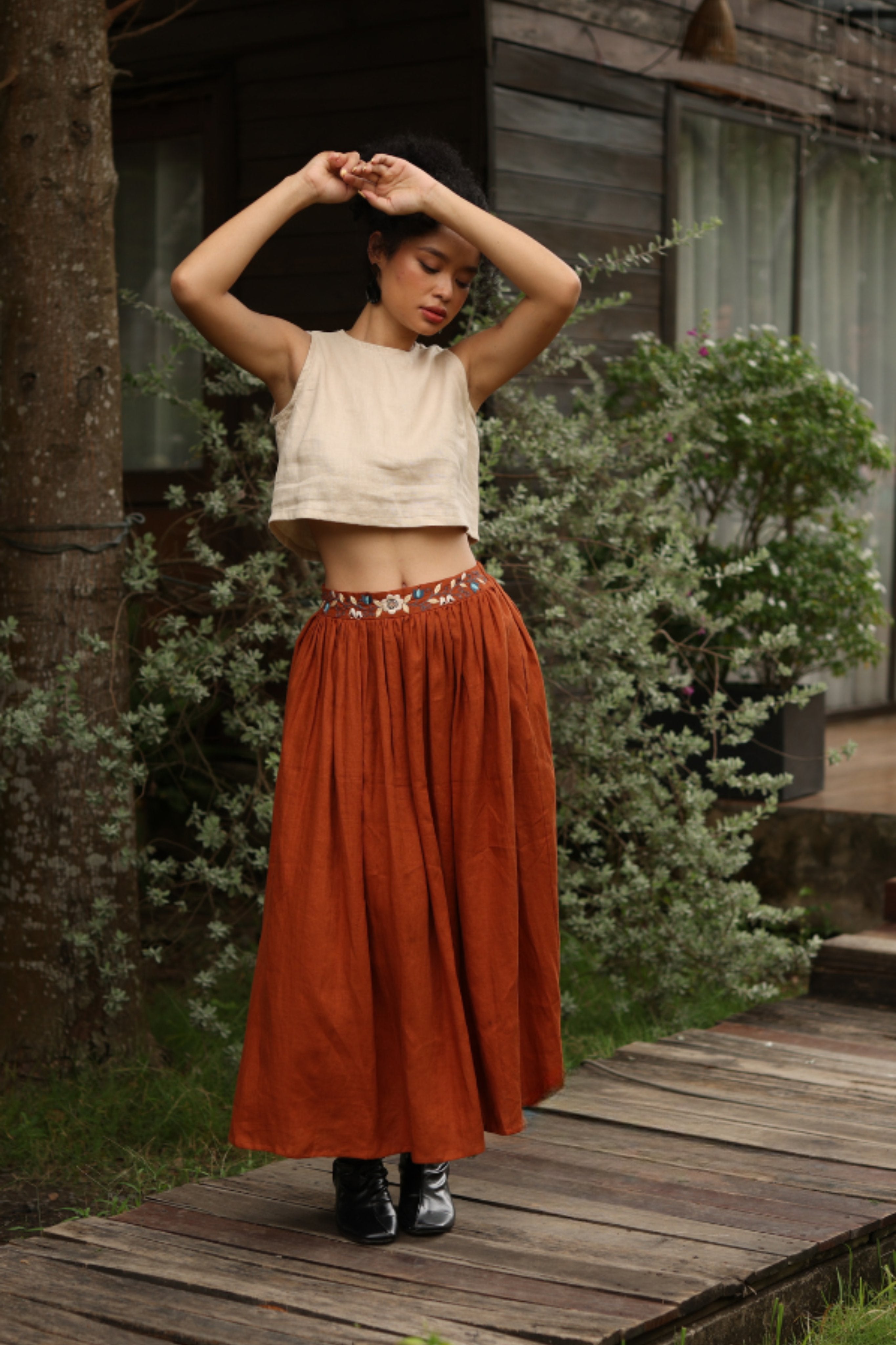 Woman in a white top and rust-colored skirt standing on a wooden path with greenery around.