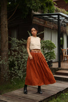 Woman in a white top and rust-colored skirt standing on a wooden path with greenery and a building in the background.