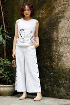 Woman wearing a sleeveless floral top and white pants against a textured wall.