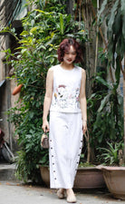 Woman in a white sleeveless top and pants standing among green plants.