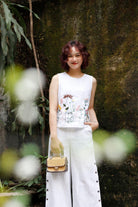 Woman in a white outfit with floral design holding a woven handbag, standing against a textured wall.