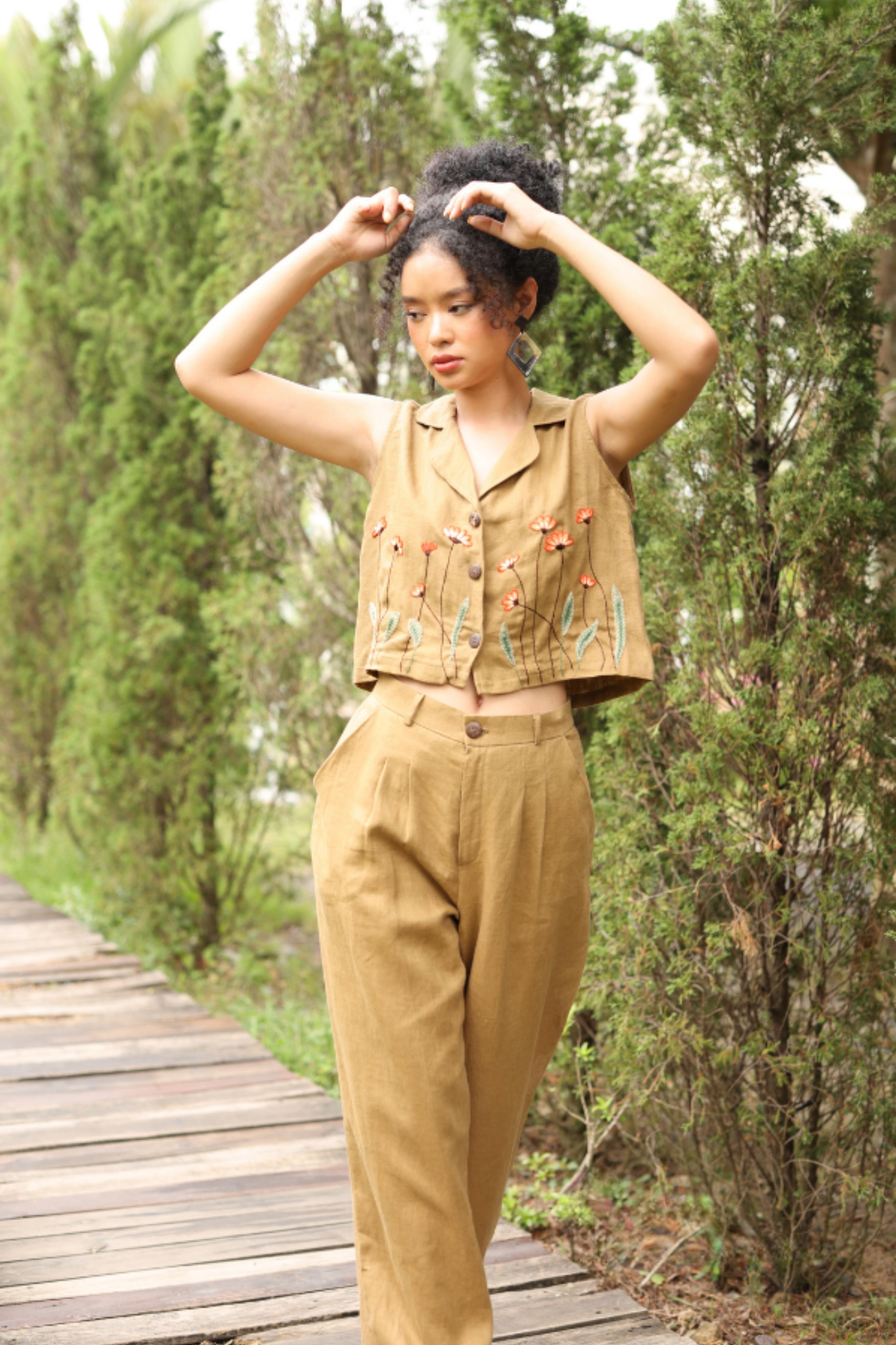 Woman in a vest and pants standing on a wooden path with greenery around.