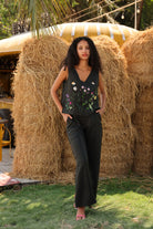 Woman standing in front of hay bales wearing a black sleeveless top with floral embroidery and black pants.