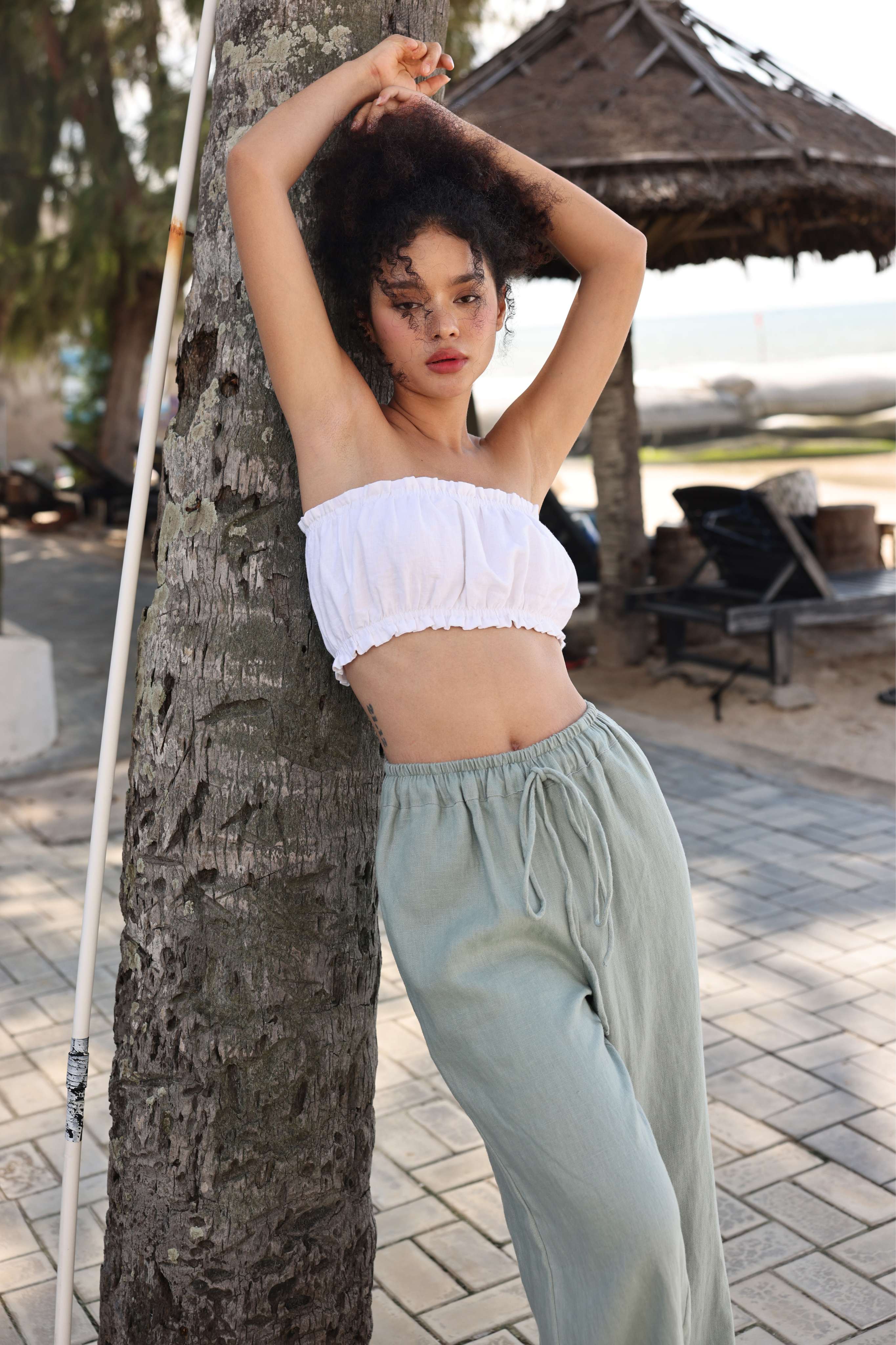 Woman in a white top and light green pants leaning against a tree in a casual setting.