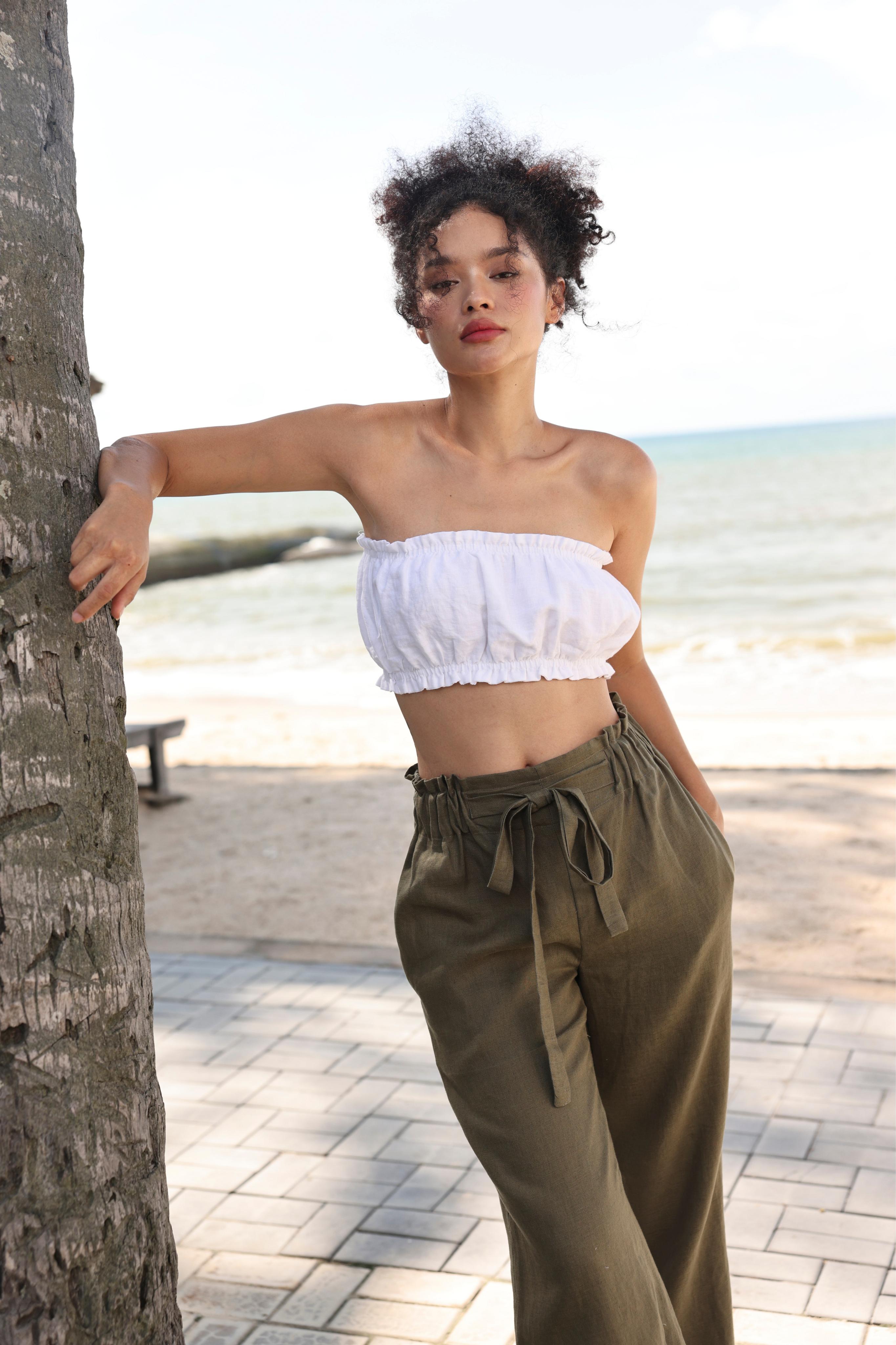 Woman in white top and olive green pants leaning against a palm tree on a beach.