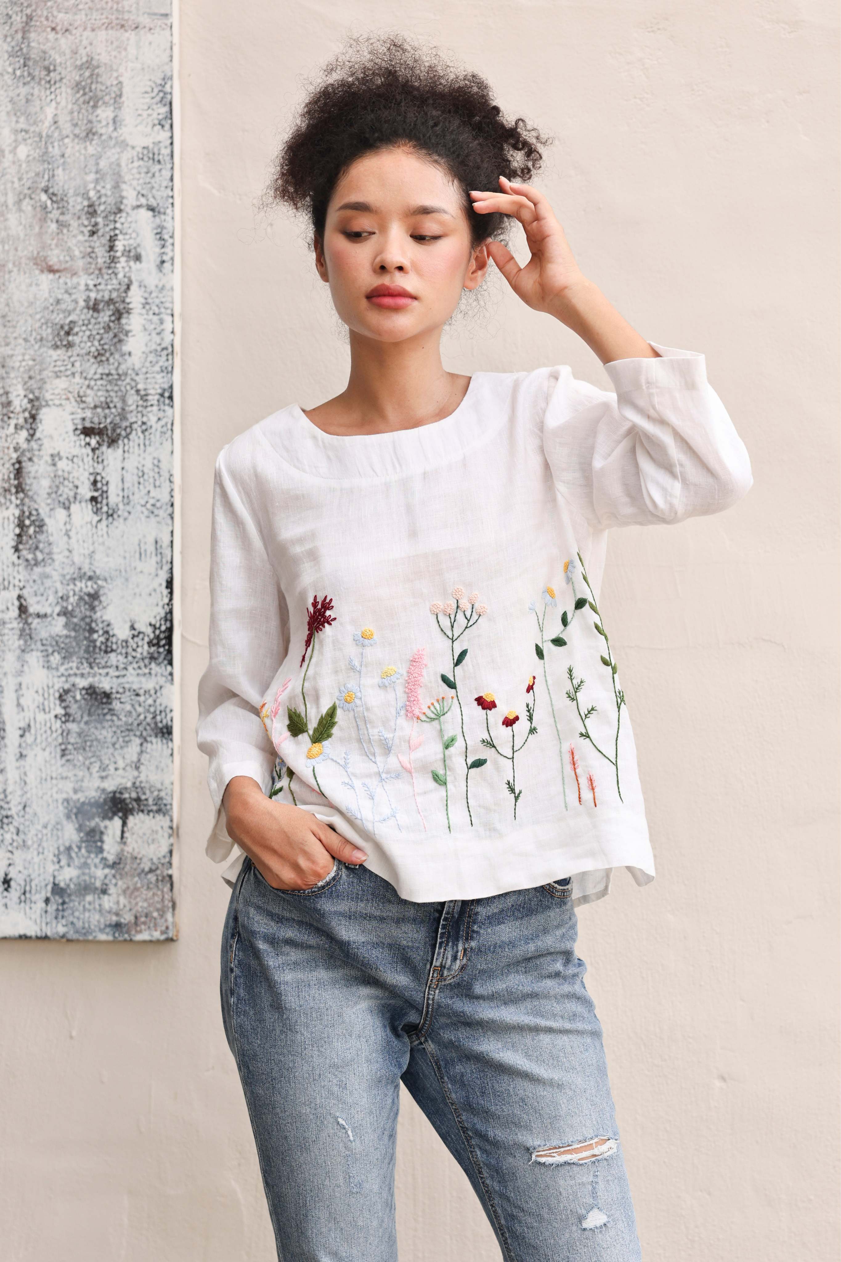 Woman wearing a white blouse with floral embroidery and blue jeans against a neutral background