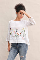 Woman wearing a white blouse with floral embroidery and blue jeans against a beige background