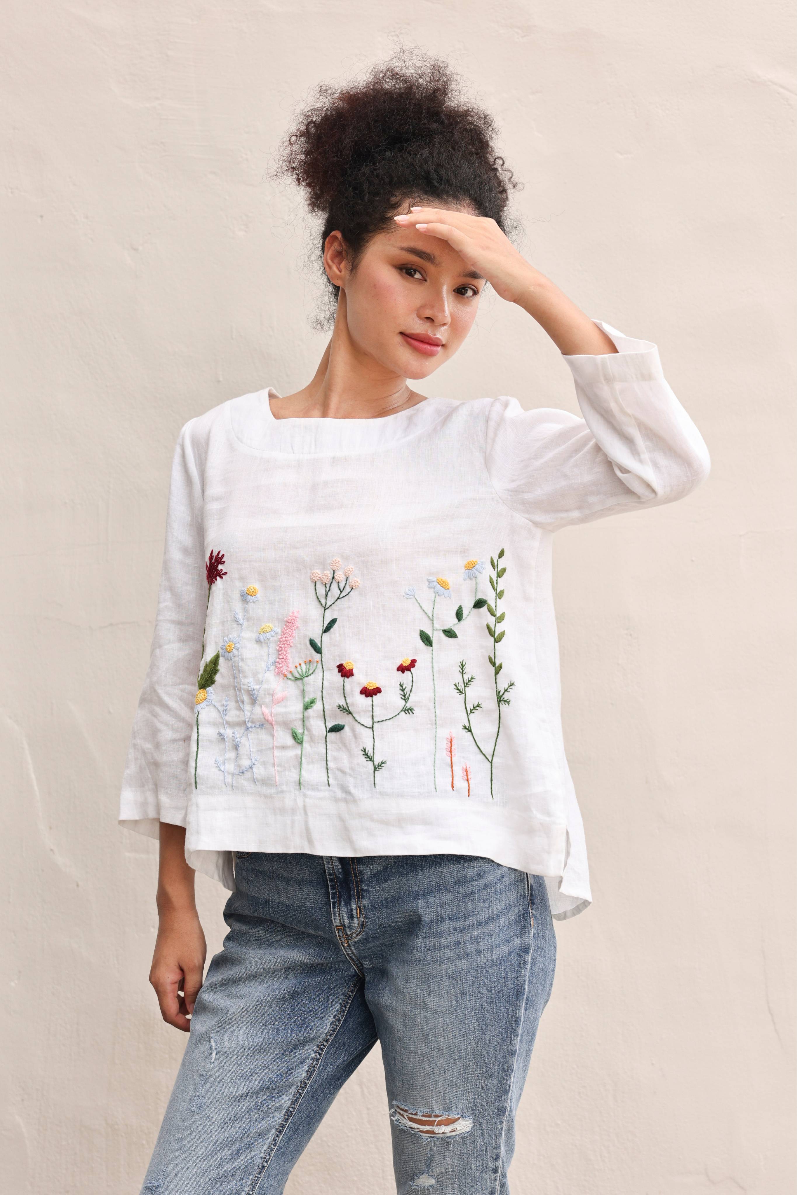 Woman wearing a white blouse with floral embroidery and blue jeans against a beige background