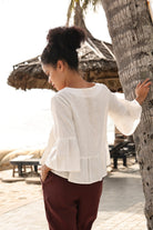 Woman in a white blouse and maroon pants standing by a tree in a tropical setting.