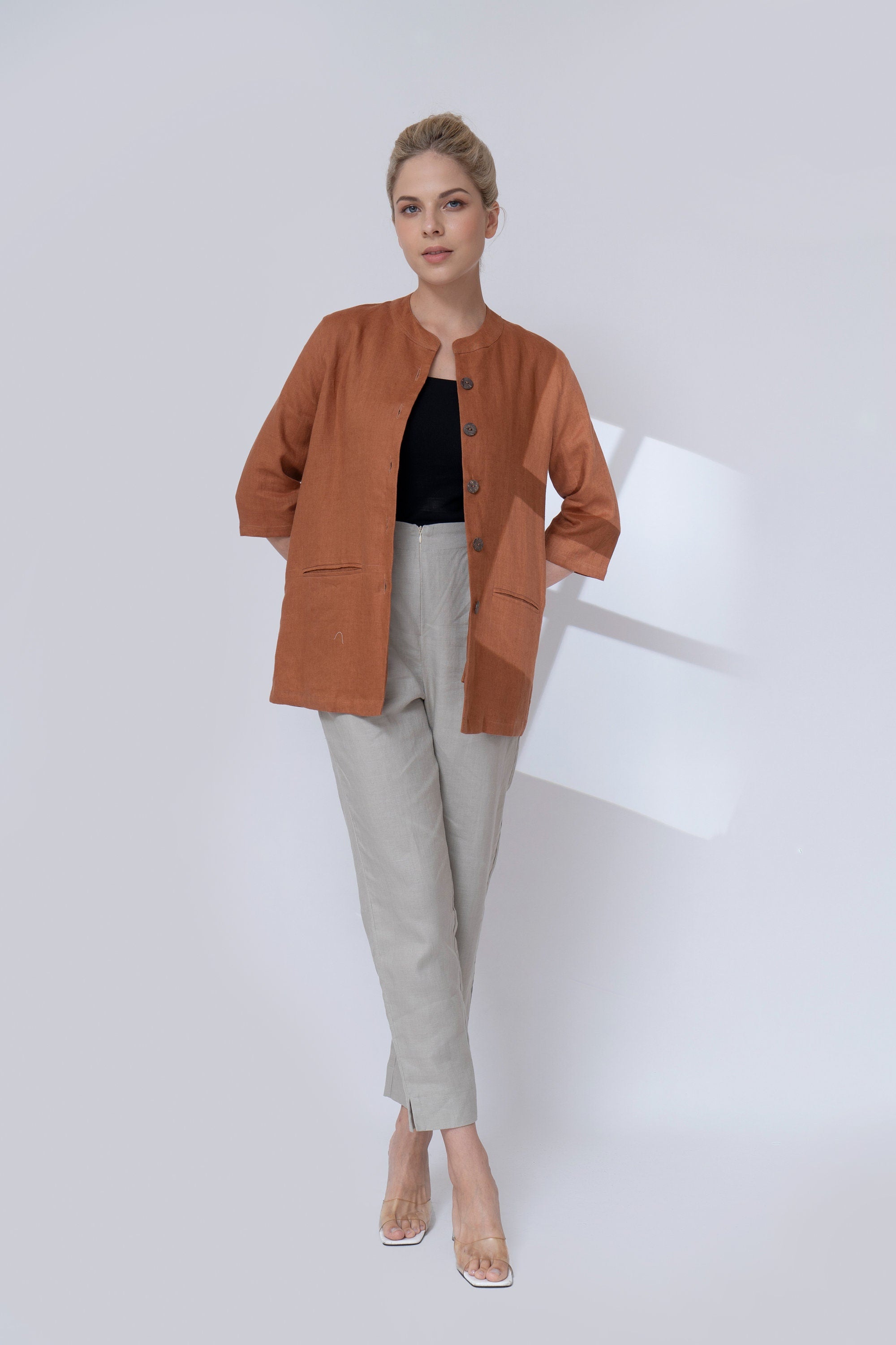 Woman wearing a brown jacket, black top, and light gray pants on a white background