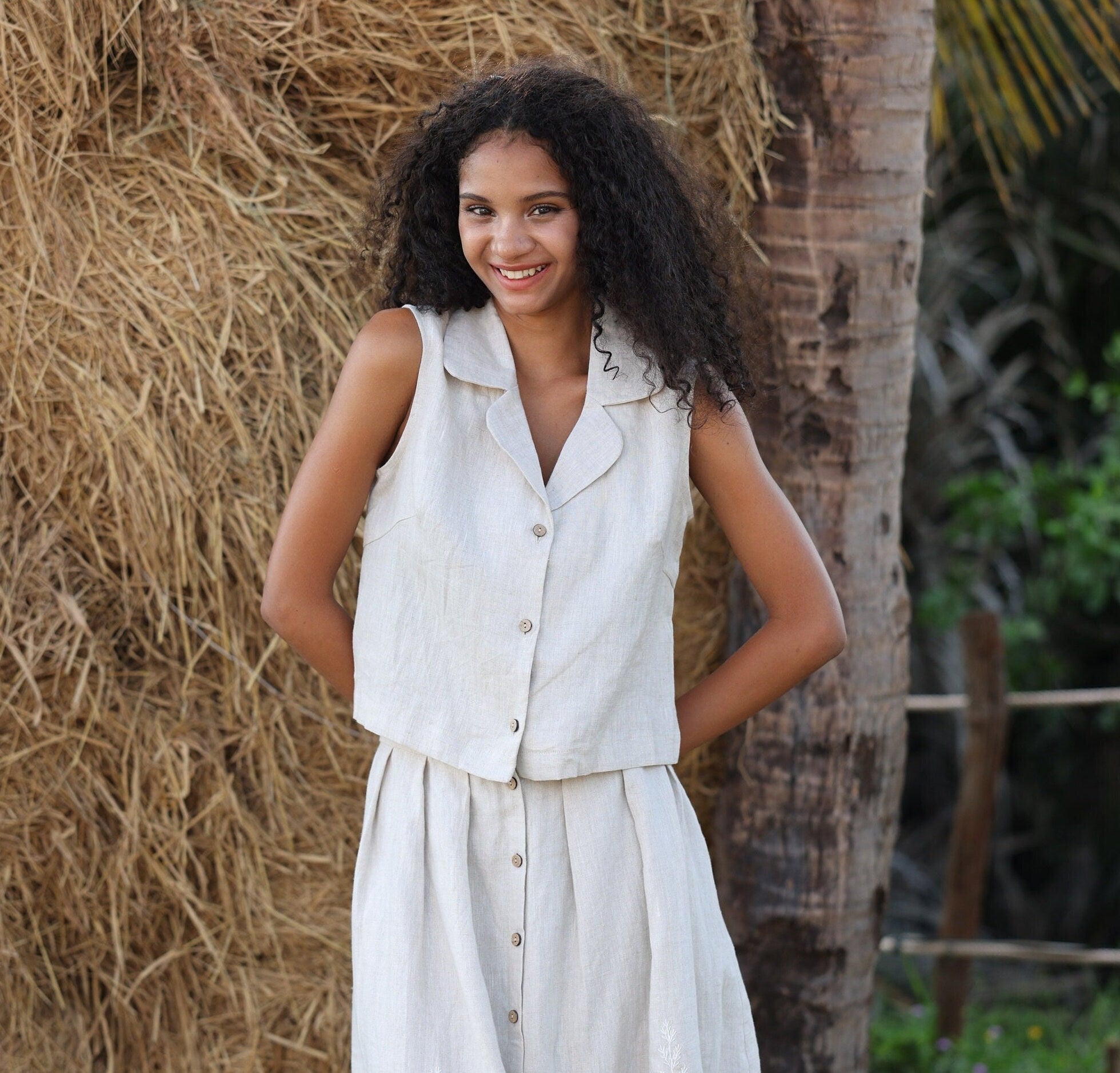Woman in a light-colored sleeveless vest top standing against a natural background with hay and a tree.