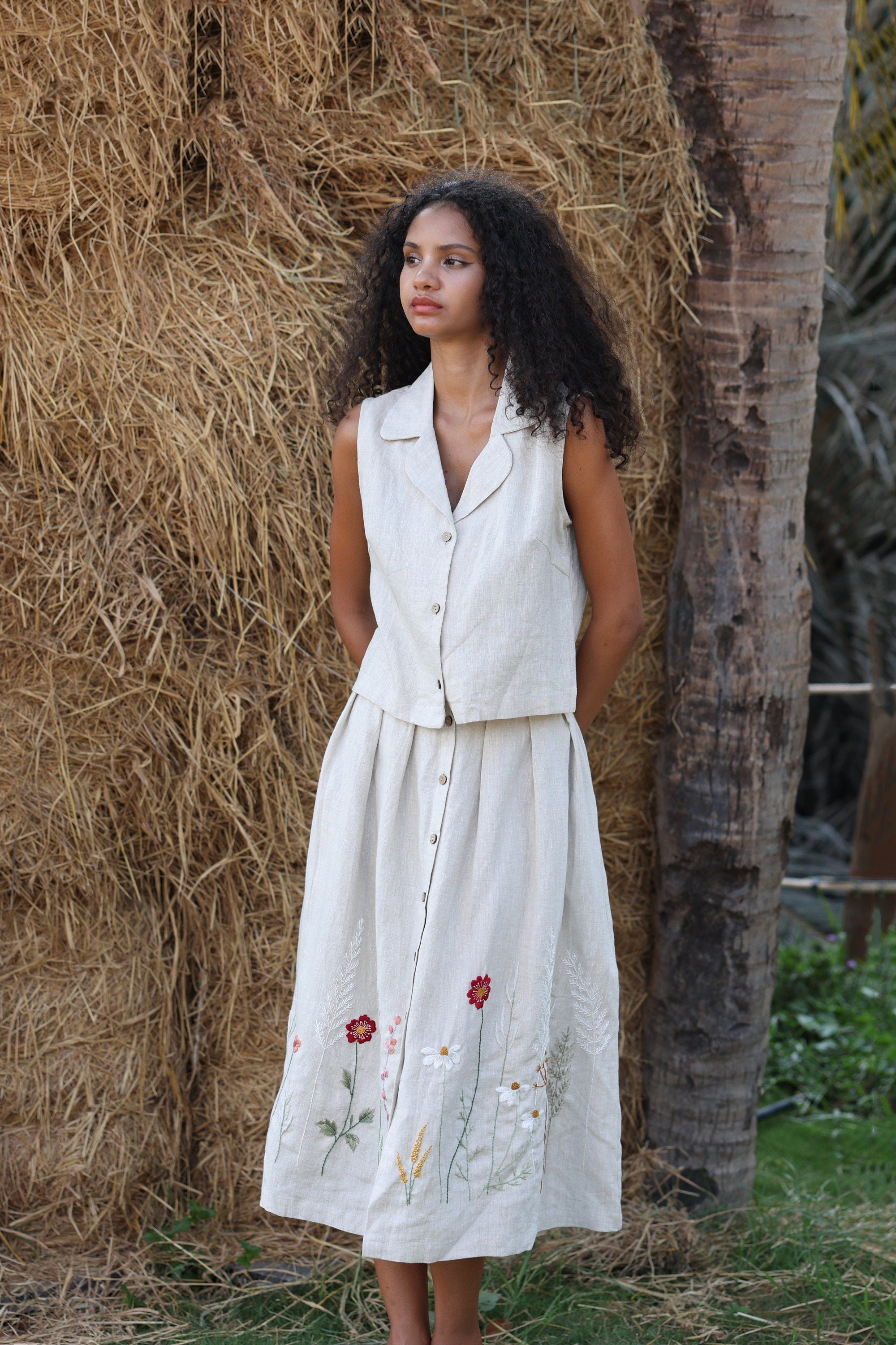 Woman wearing a top and a floral-embroidered skirt standing next to a stack of hay.