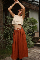 Woman in a beige top and rust-colored skirt standing outdoors with plants and a building in the background.