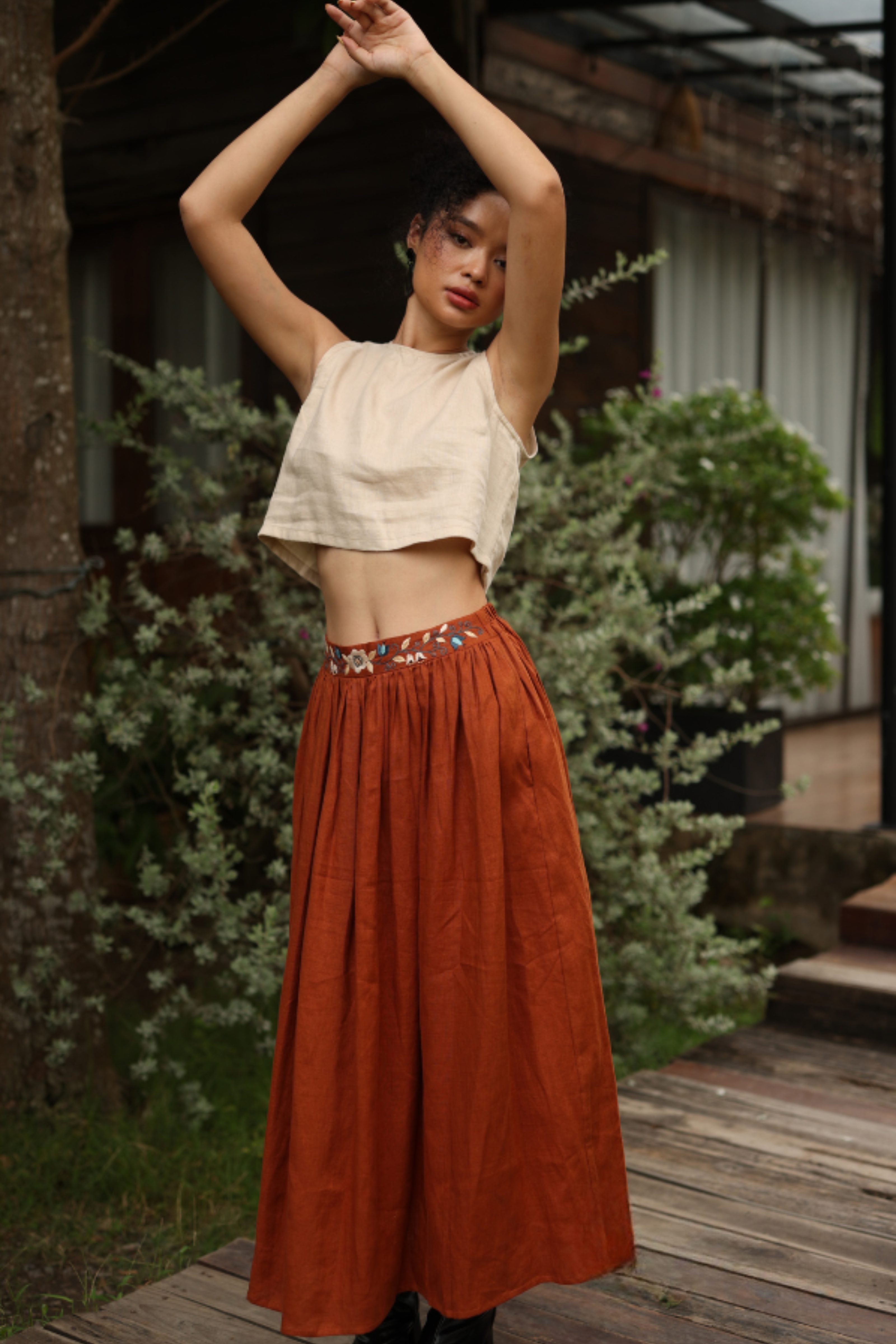 Woman in a beige top and rust-colored skirt standing outdoors with plants and a building in the background.