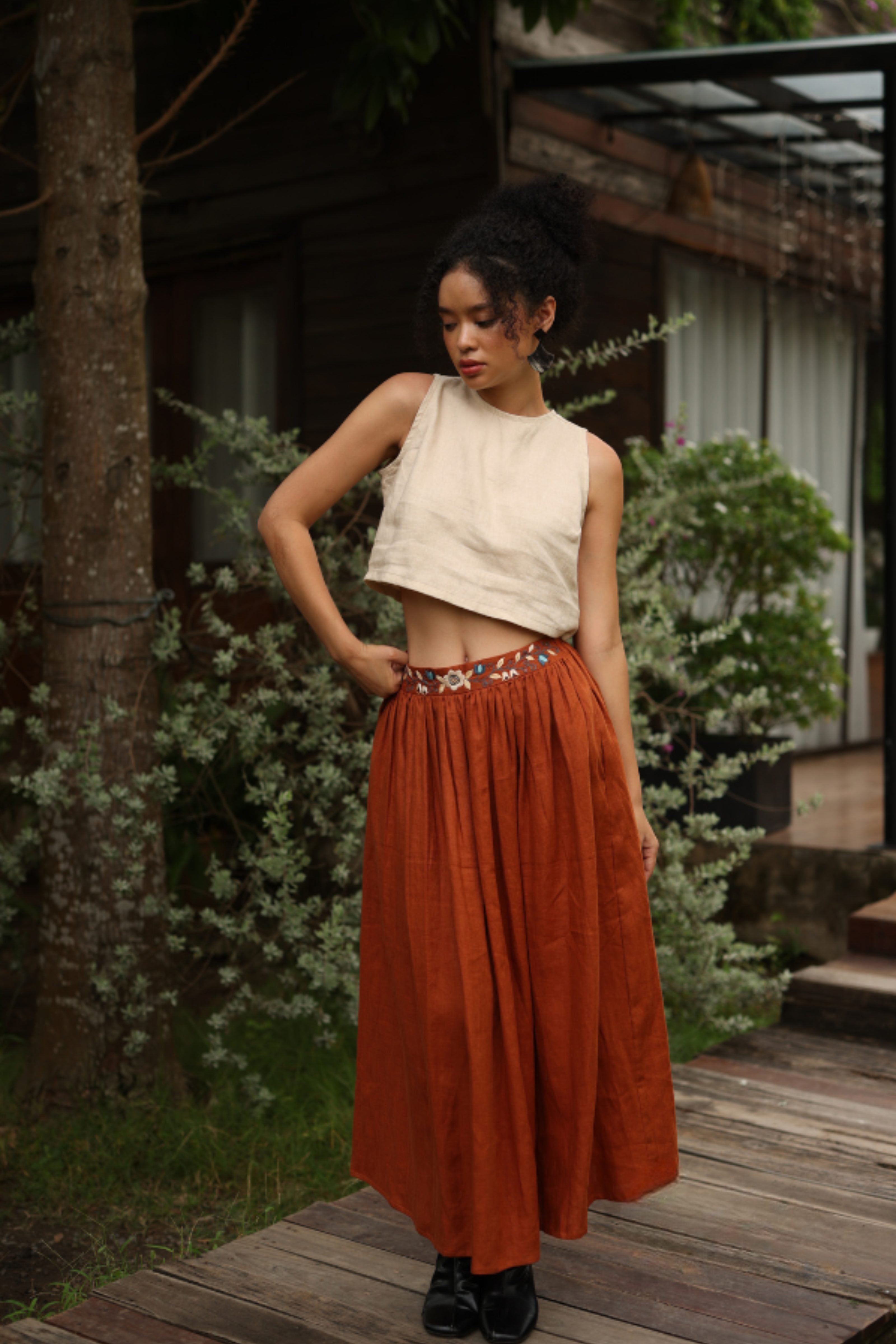 Woman wearing a white top and rust-colored skirt standing outdoors with greenery in the background