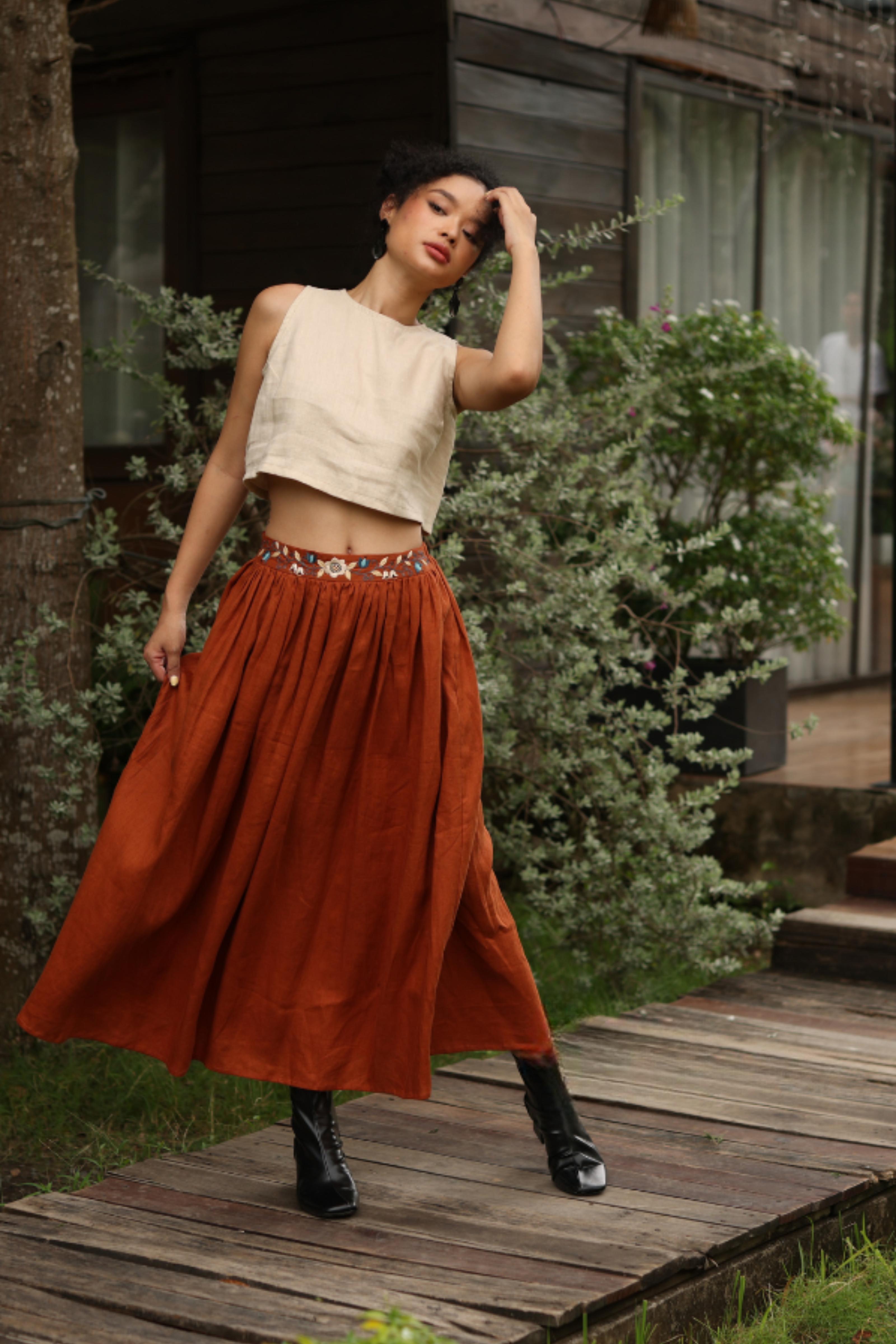 Woman in a beige top and rust-colored skirt standing on a wooden path with greenery in the background