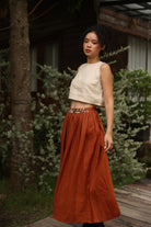 Woman wearing a white top and rust-colored skirt standing outdoors with greenery in the background