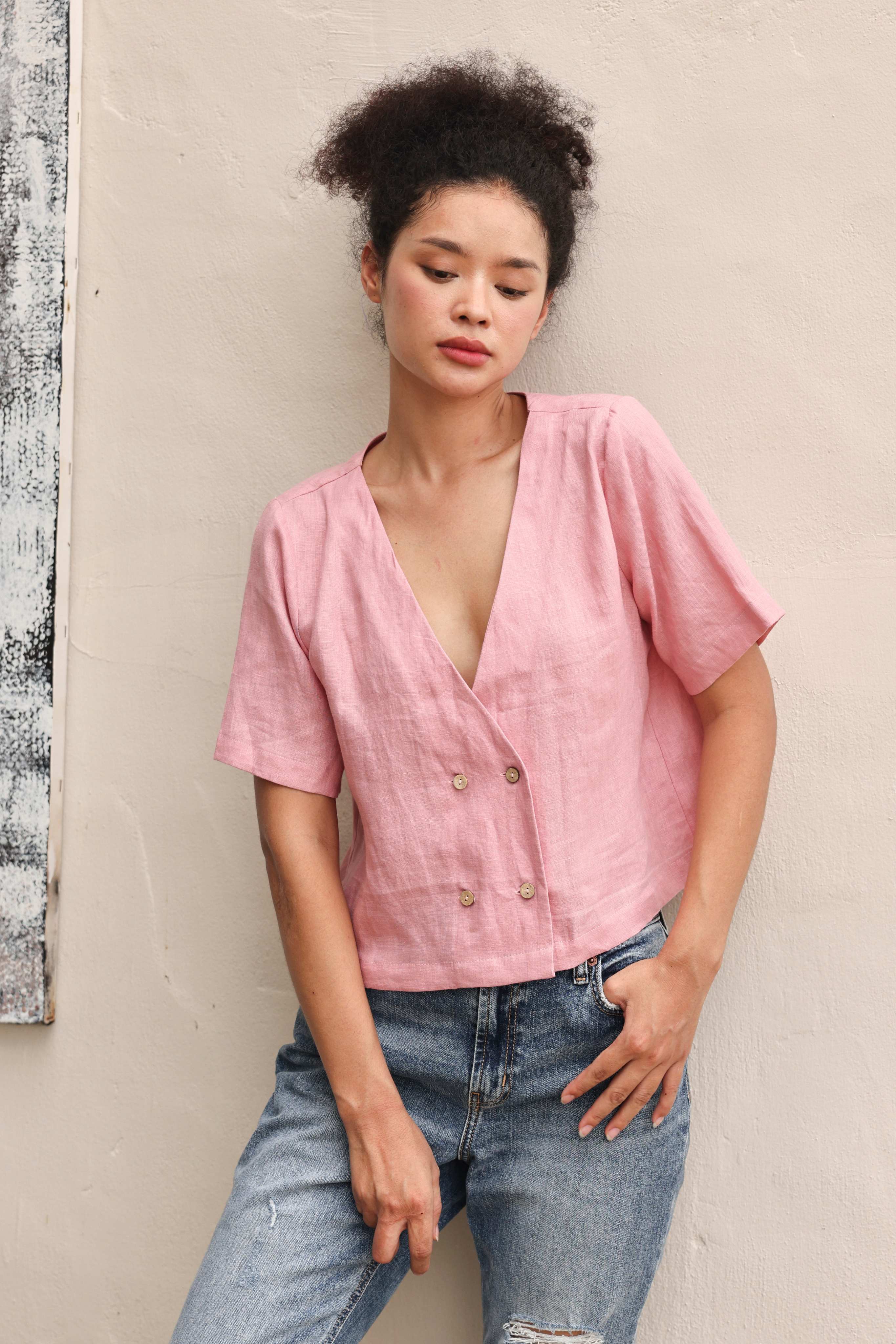 Woman wearing a pink blouse with buttons and blue jeans against a beige wall.