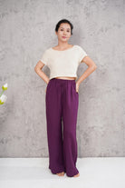 Woman wearing a white top and purple pants against a gray concrete wall.