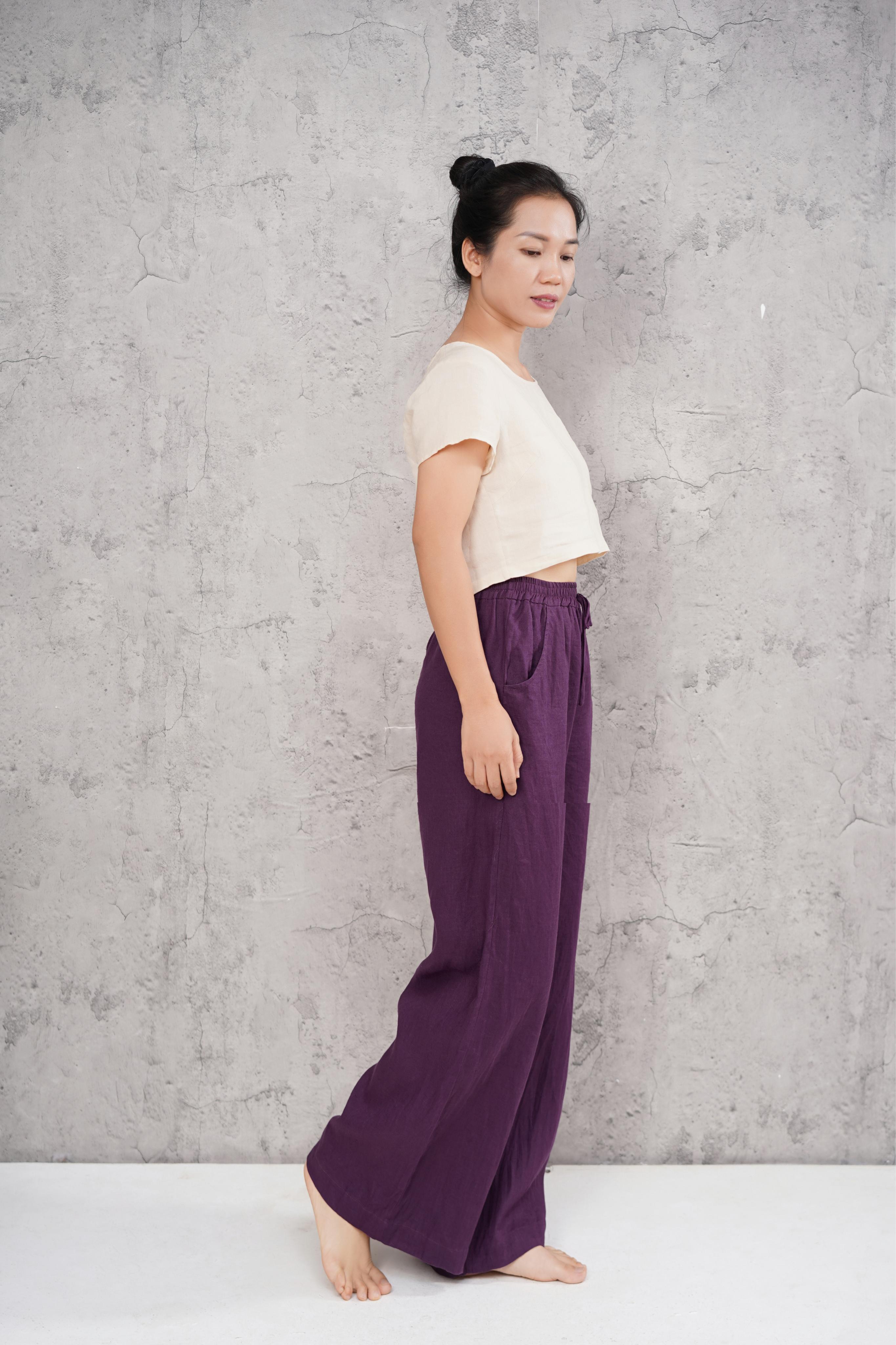 Woman wearing a white top and purple pants against a gray concrete wall.