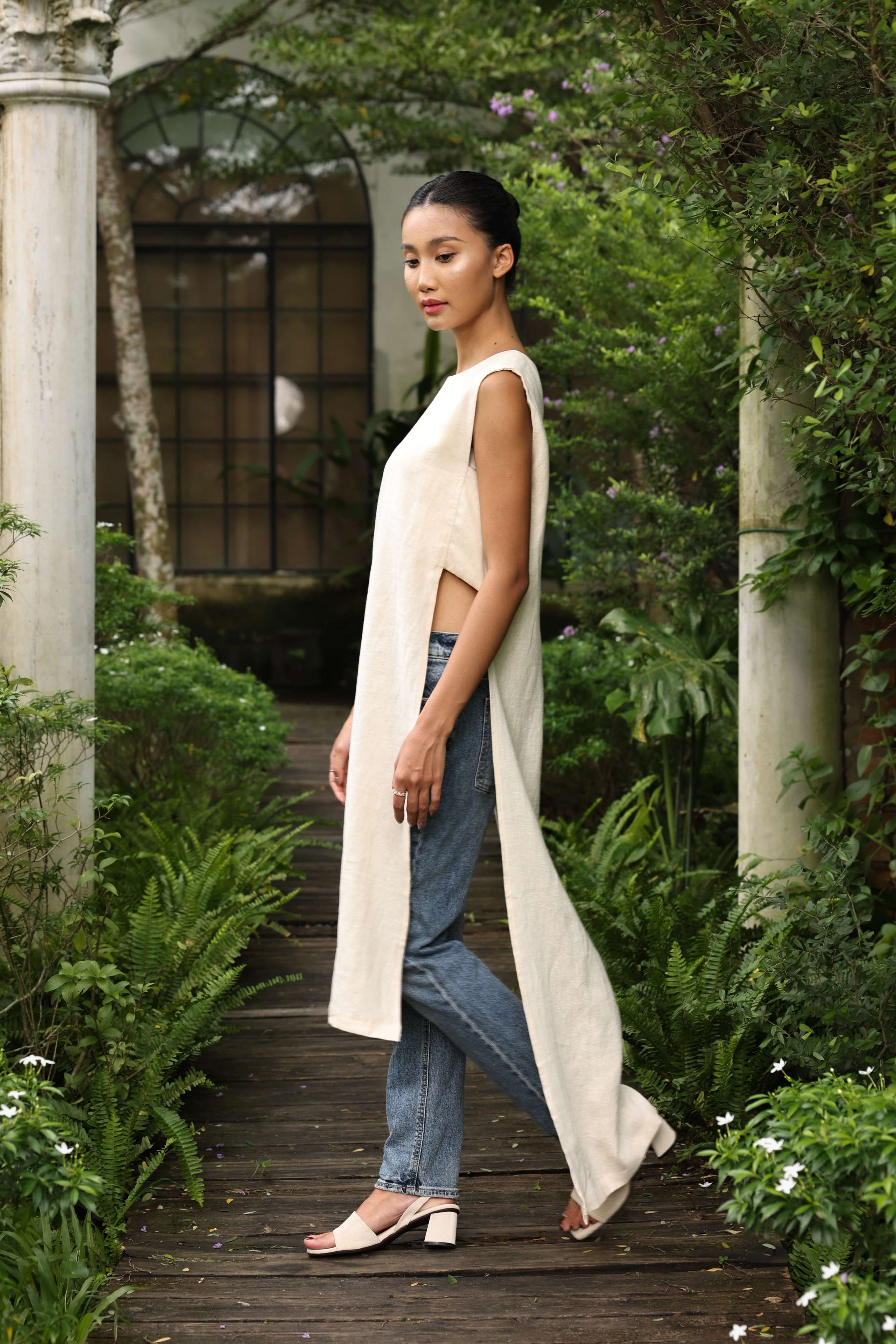 Woman walking through a garden wearing a long beige garment over a sleeveless top and jeans.