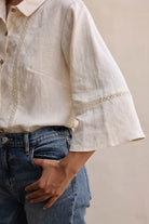 Person wearing a beige linen shirt with lace trim and blue jeans against a neutral background