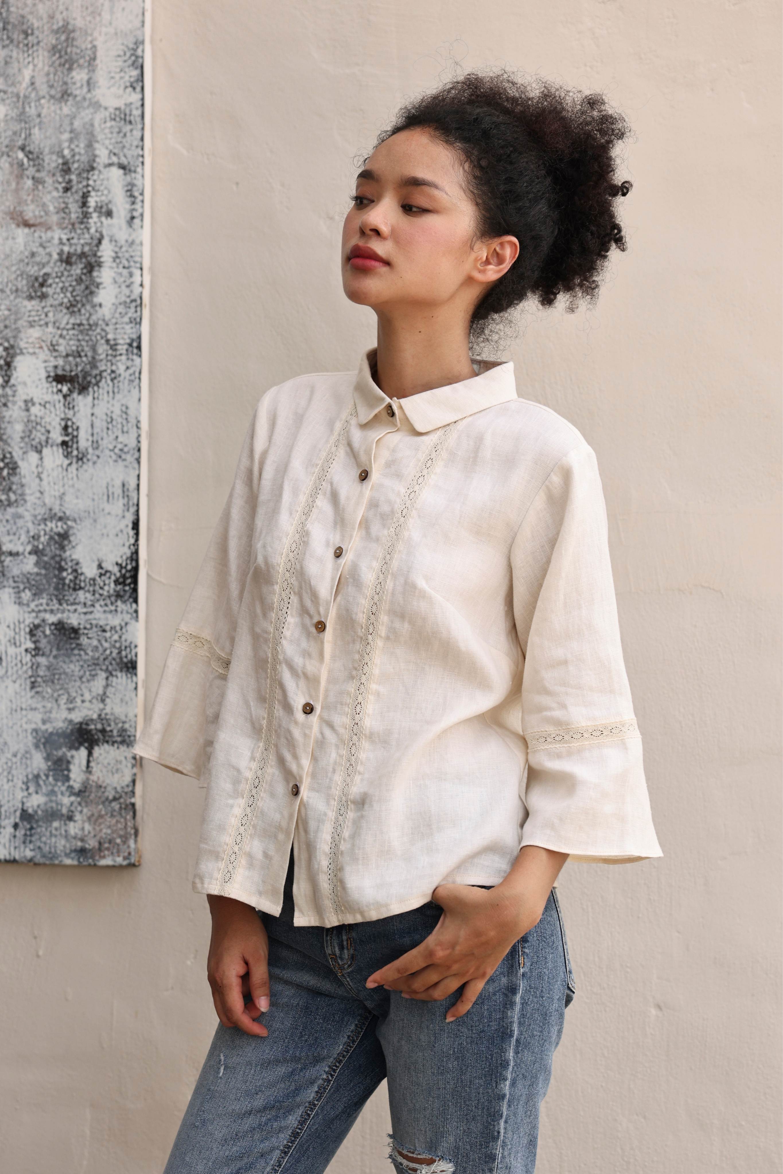 Woman wearing a beige button-up shirt and jeans against a neutral wall.
