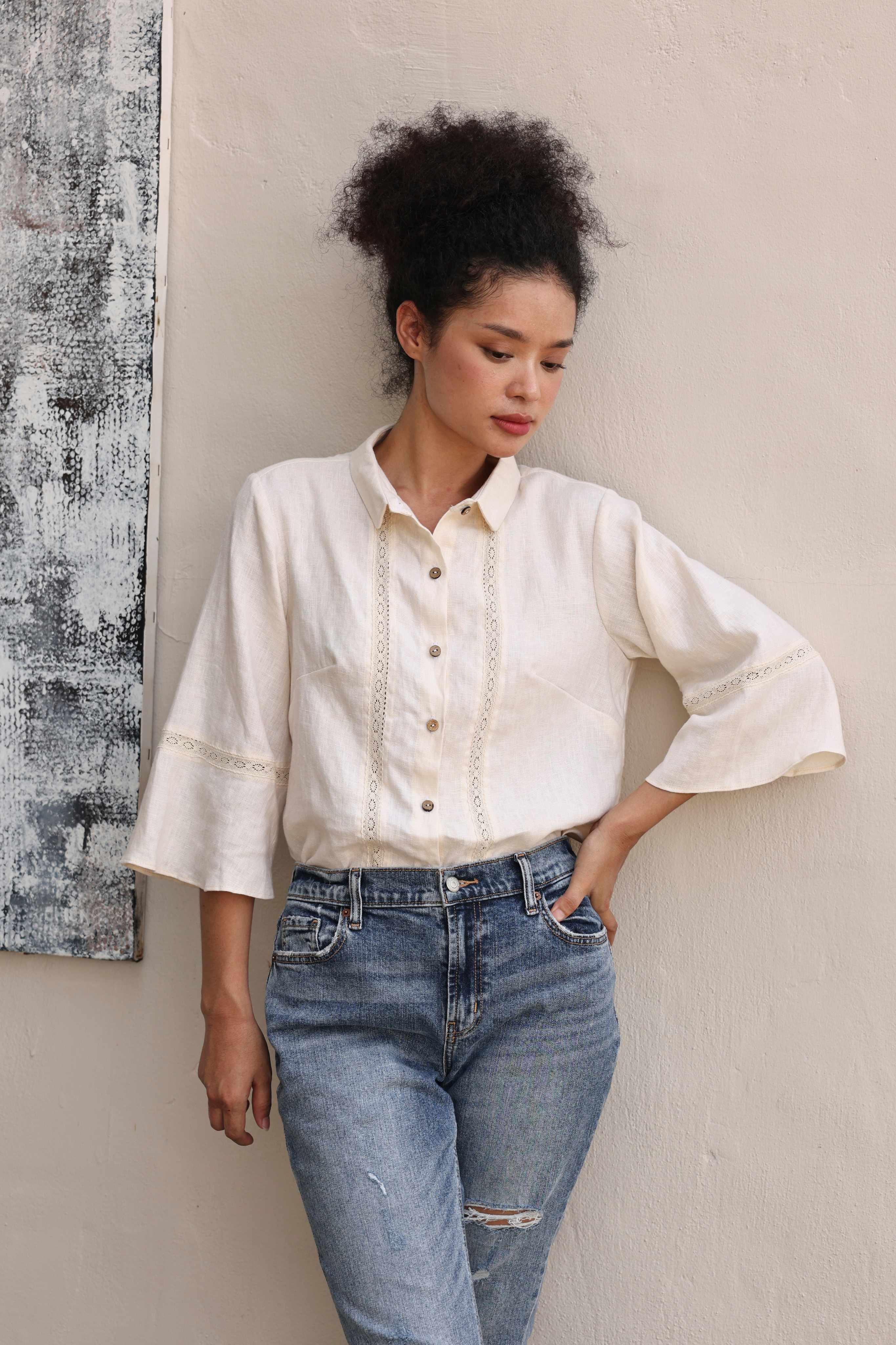Woman wearing a beige shirt and blue jeans against a neutral wall.