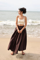 Woman standing on a beach wearing a beige top and brown skirt.
